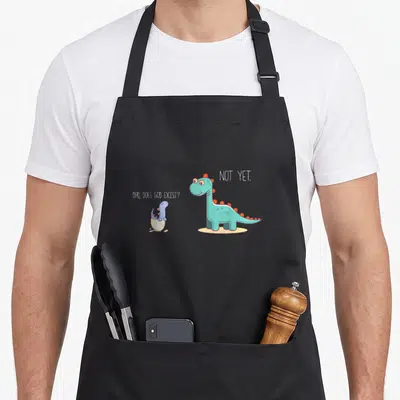 Doe dinosaur apron - Teal dinosaur answers Not Yet to hatchling asking if God exists — shown on a black cotton canvas apron against a white background