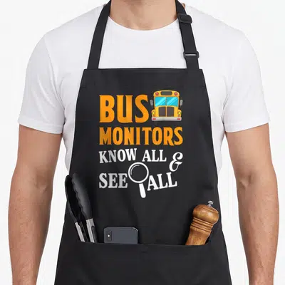 See omnibus apron - Orange BUS text, yellow school bus, white KNOW ALL SEE ALL with magnifying glass — shown on a black cotton canvas apron against a white background