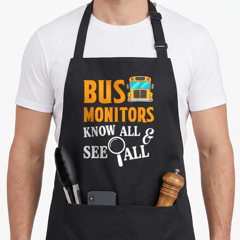 See omnibus apron - Orange BUS text, yellow school bus, white KNOW ALL SEE ALL with magnifying glass — shown on a black cotton canvas apron against a white background