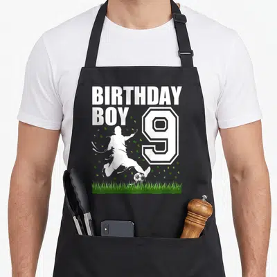 Theme 9th birthday apron - BIRTHDAY BOY text next to number 9 and soccer player silhouette kicking ball over green grass — shown on a black cotton canvas apron against a white background