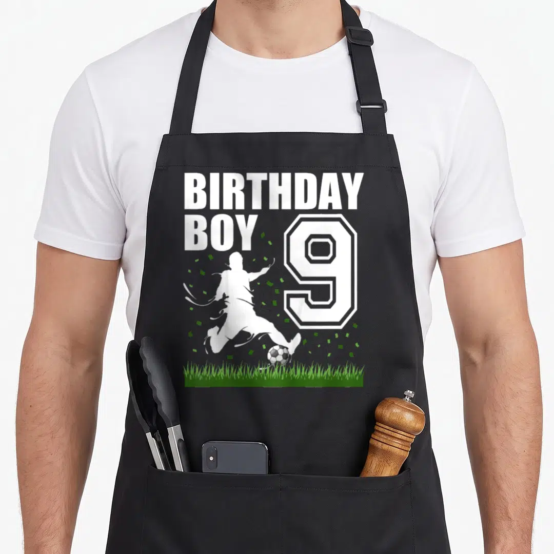 Theme 9th birthday apron - BIRTHDAY BOY text next to number 9 and soccer player silhouette kicking ball over green grass — shown on a black cotton canvas apron against a white background