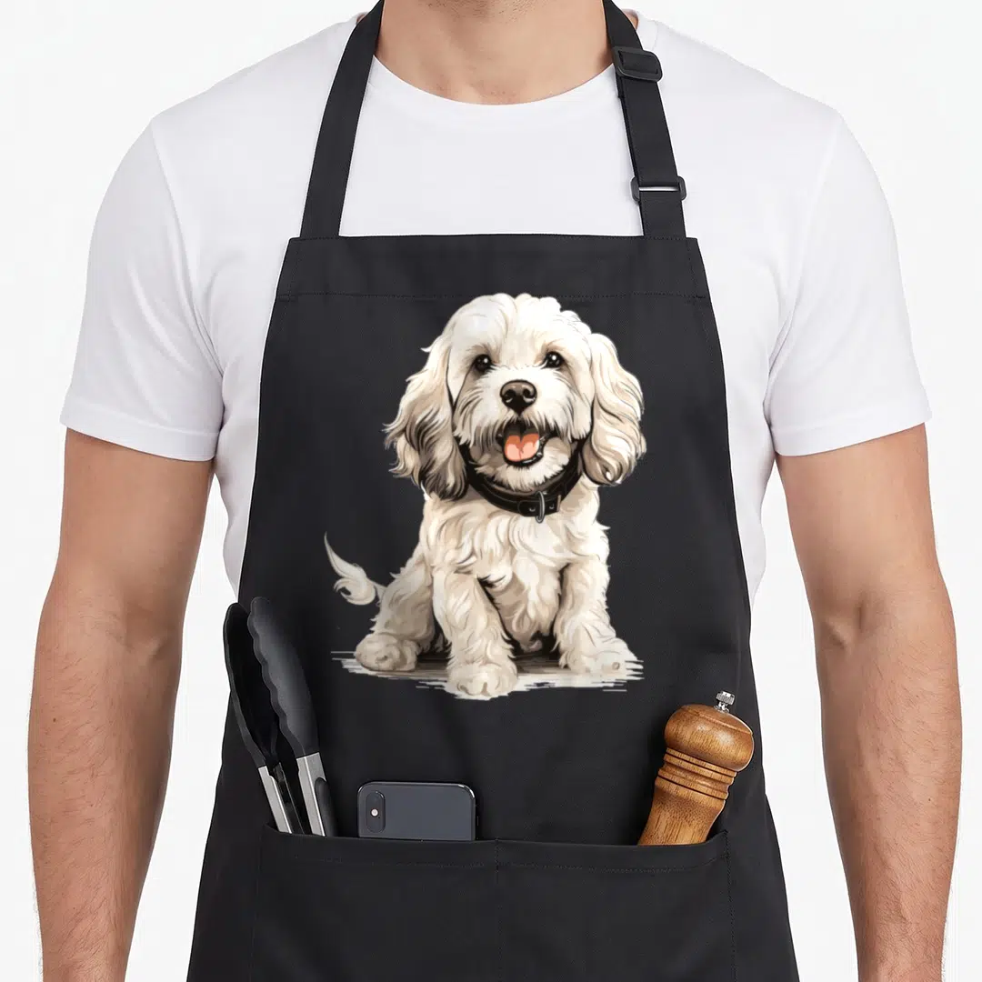 Breed cockapoo apron - Cartoon cream dog sitting facing forward with black collar and pink tongue — shown on a black cotton canvas apron against a white background