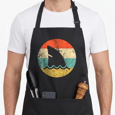 Fin shark apron - Grey shark fin and waves inside circle of red cream teal yellow bands — shown on a black cotton canvas apron against a white background