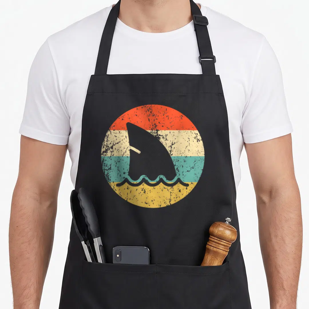 Fin shark apron - Grey shark fin and waves inside circle of red cream teal yellow bands — shown on a black cotton canvas apron against a white background