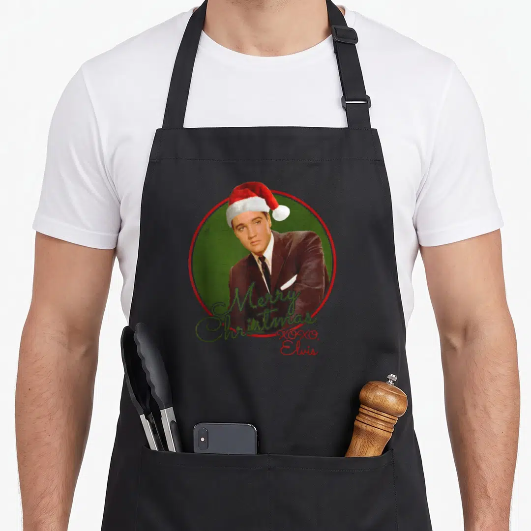 Elvis merry christmas apron - Elvis photo in Santa hat inside green circle with Merry Christmas XOXO script text — shown on a black cotton canvas apron against a white background