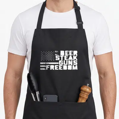 Steak american flag apron - Distressed white text BEER STEAK GUNS FREEDOM next to white stars and stripes on grey — shown on a black cotton canvas apron against a white background