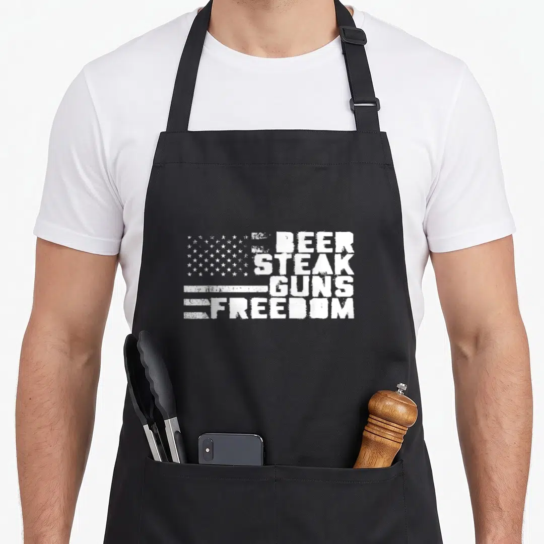 Steak american flag apron - Distressed white text BEER STEAK GUNS FREEDOM next to white stars and stripes on grey — shown on a black cotton canvas apron against a white background