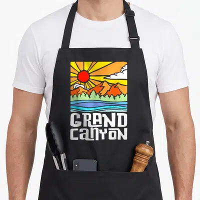 Grand artistic apron - Retro landscape with red sun, orange mountains, green trees, blue water, and white GRAND CANYON — shown on a black cotton canvas apron against a white background