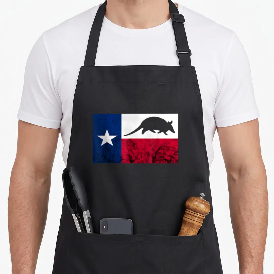 Cowboy texas flag apron - Grey armadillo silhouette over distressed blue white and red Texas flag with single white star — shown on a black cotton canvas apron against a white background