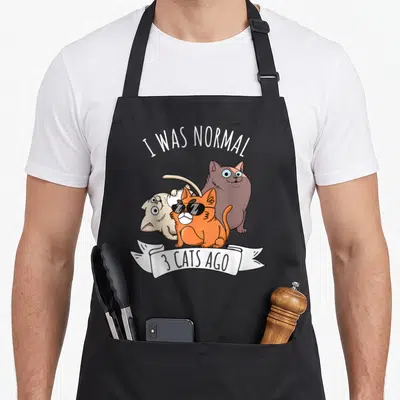 Normal crazy cat lady apron - I Was Normal 3 Cats Ago text over orange, brown, beige cartoon cats with white ribbon — shown on a black cotton canvas apron against a white background