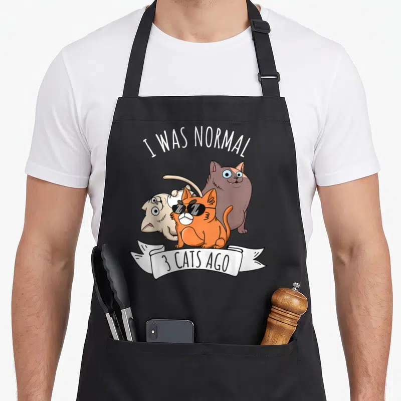 Normal crazy cat lady apron - I Was Normal 3 Cats Ago text over orange, brown, beige cartoon cats with white ribbon — shown on a black cotton canvas apron against a white background