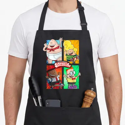 Underpant friends apron - Captain Underpants four panel grid with red yellow blue green backgrounds and central title — shown on a black cotton canvas apron against a white background