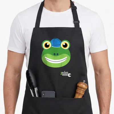 Gecko garage apron - Green cartoon gecko head in blue cap with gecko's GARAGE text and white wrench logo — shown on a black cotton canvas apron against a white background