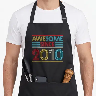 Vintage 13th age birthday apron - Stacked striped text reading AWESOME SINCE 2010 in yellow red white and cyan — shown on a black cotton canvas apron against a white background