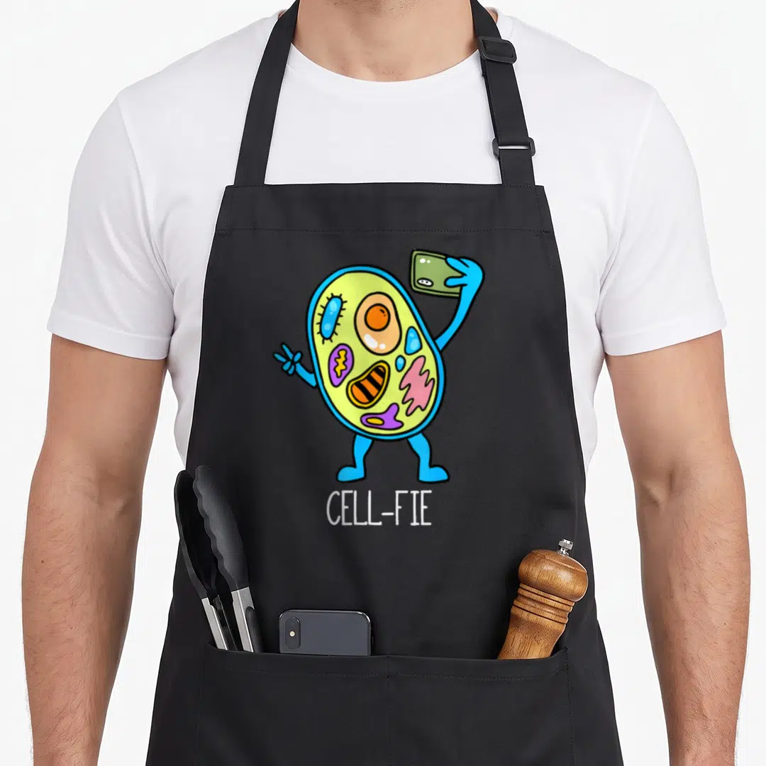 Cellfie teacher apron - Cartoon cell with blue arms holding a phone, lime green body with colorful organelles, white — shown on a black cotton canvas apron against a white background