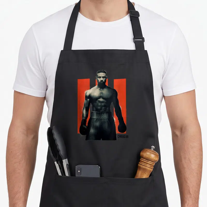 Son apollo apron - Black and white boxer photo between two red vertical bars with CREED III text — shown on a black cotton canvas apron against a white background