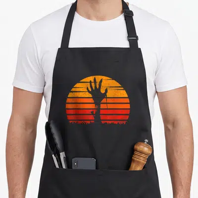 Sunset halloween apron - Grey hand silhouette rising from grass against striped orange and red retro sun — shown on a black cotton canvas apron against a white background