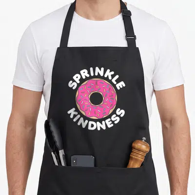 Strawberry sprinkle apron - Pink frosted donut with SPRINKLE KINDNESS text in white and tan — shown on a black cotton canvas apron against a white background