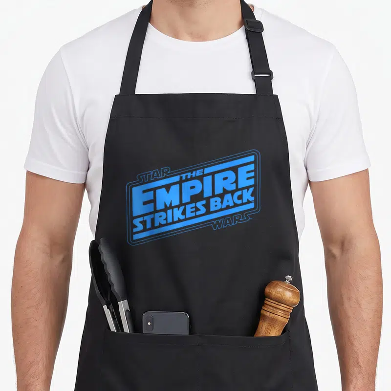 Empire logo officially apron - Blue stacked text reading Star Wars The Empire Strikes Back inside a rectangular frame — shown on a black cotton canvas apron against a white background