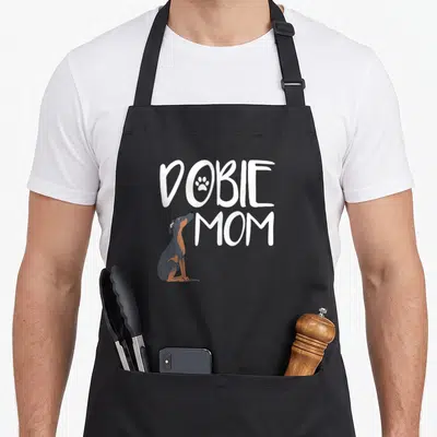 Puppy pet dog apron - White DOBIE MOM text with paw print and sitting grey tan dog — shown on a black cotton canvas apron against a white background