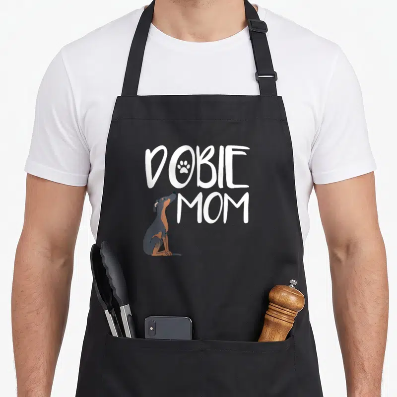 Puppy pet dog apron - White DOBIE MOM text with paw print and sitting grey tan dog — shown on a black cotton canvas apron against a white background