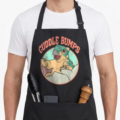 Jurassic bump apron - Cartoon dinosaur inside teal circle with arched orange text CUDDLE BUMPS — shown on a black cotton canvas apron against a white background