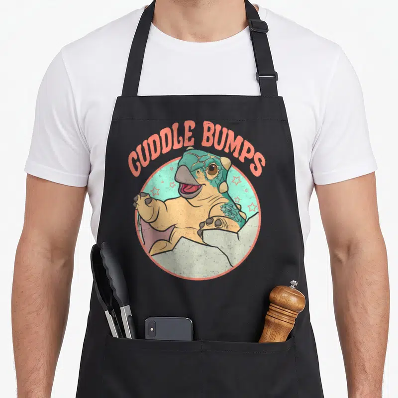 Jurassic bump apron - Cartoon dinosaur inside teal circle with arched orange text CUDDLE BUMPS — shown on a black cotton canvas apron against a white background