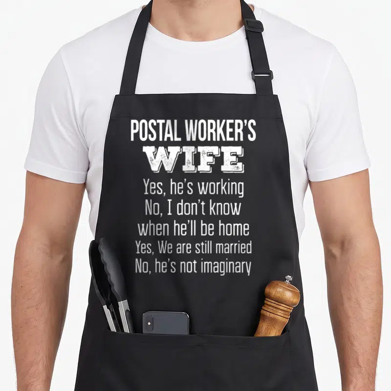 Postal wife apron - White stacked text reading Postal Workers Wife Yes he is working No I do not know when he will be — shown on a black cotton canvas apron against a white background