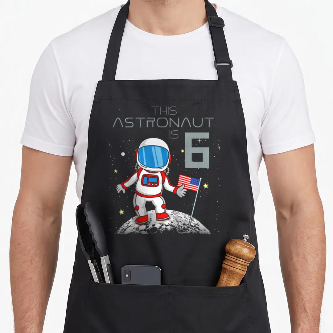 6th geek apron - Cartoon astronaut holding flag on moon with This Astronaut Is 6 text — shown on a black cotton canvas apron against a white background