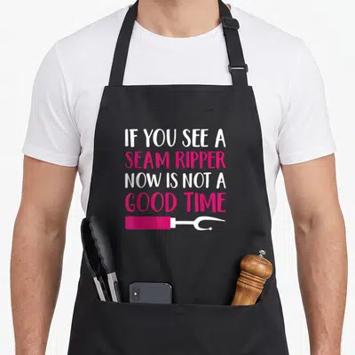 Sewing dressmaking apron - Four lines of white and pink text reading if you see a seam ripper above a pink tool — shown on a black cotton canvas apron against a white background