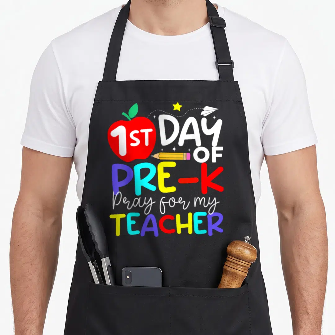 Pray teacher apron - Red apple with 1ST text, pencil, star, paper plane, and multi-colored PRE-K TEACHER lettering — shown on a black cotton canvas apron against a white background