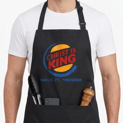 Christ god king of the grill apron - Stacked red text in blue and orange circle with blue caption below — shown on a black cotton canvas apron against a white background