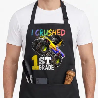 Cap truck apron - Monster truck with graduation cap and mud splatters reading I CRUSHED 1ST GRADE — shown on a black cotton canvas apron against a white background