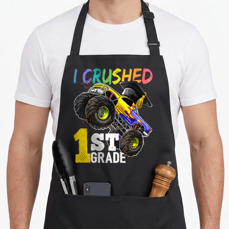 Cap truck apron - Monster truck with graduation cap and mud splatters reading I CRUSHED 1ST GRADE — shown on a black cotton canvas apron against a white background