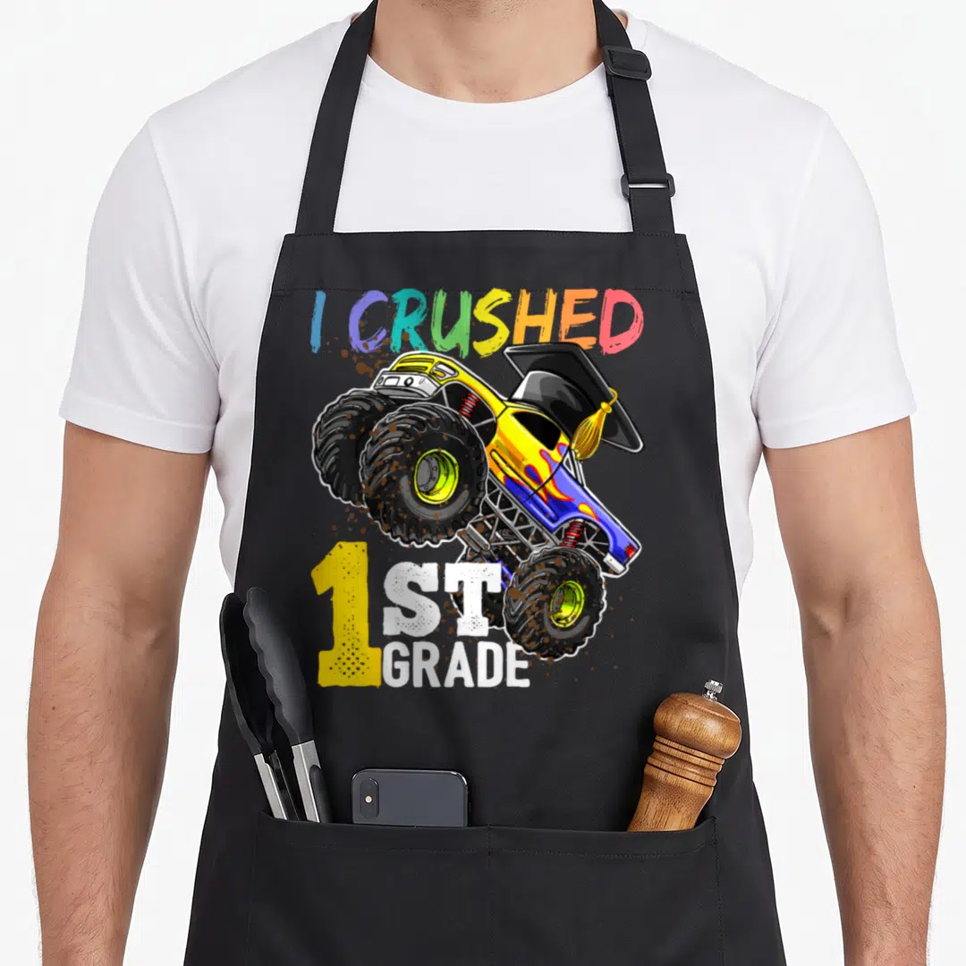 Cap truck apron - Monster truck with graduation cap and mud splatters reading I CRUSHED 1ST GRADE — shown on a black cotton canvas apron against a white background
