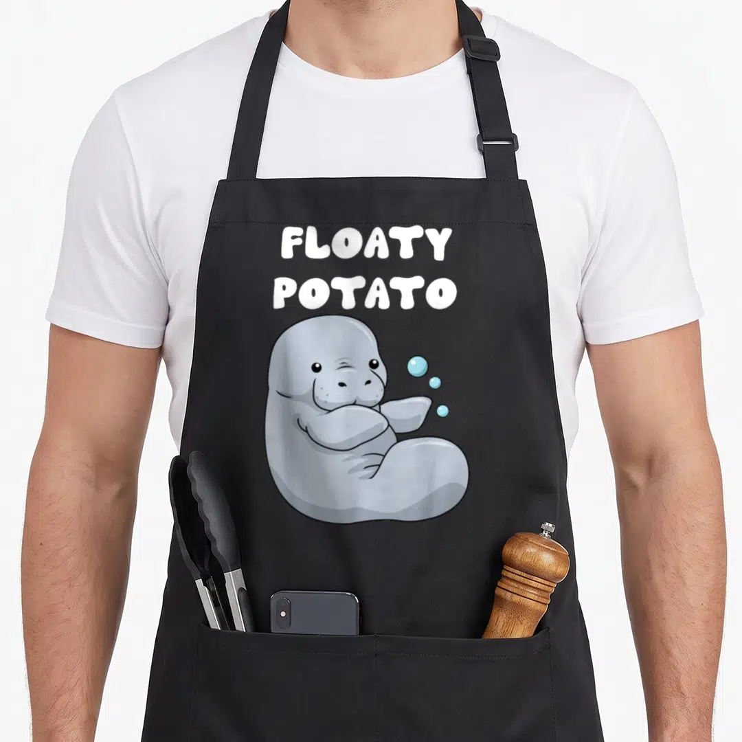 Manatee potato apron - Grey cartoon manatee winking below FLOATY POTATO text with three blue bubbles — shown on a black cotton canvas apron against a white background