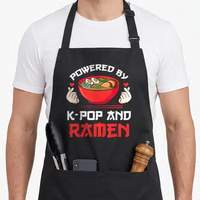 Powered k apron - Ramen bowl with two hands making finger hearts and arched text — shown on a black cotton canvas apron against a white background