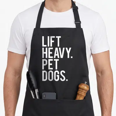 Lift heavy apron - White stacked text LIFT HEAVY PET DOGS on grey background — shown on a black cotton canvas apron against a white background
