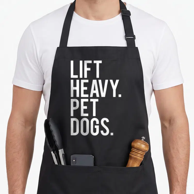 Lift heavy apron - White stacked text LIFT HEAVY PET DOGS on grey background — shown on a black cotton canvas apron against a white background