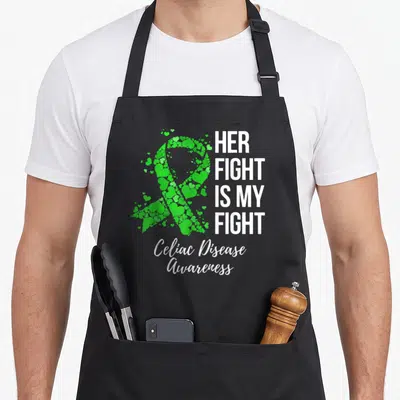 Fight celiac awareness apron - Green heart-shaped awareness ribbon next to white Her Fight Is My Fight text — shown on a black cotton canvas apron against a white background