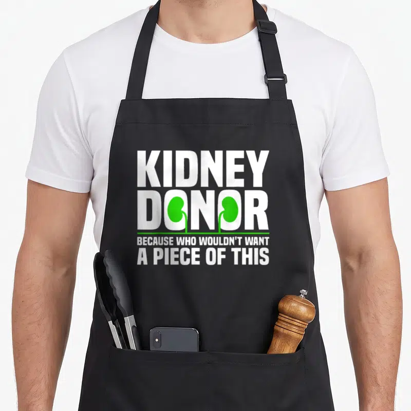 Awareness organ apron - KIDNEY DONOR text with green kidney shapes replacing letters and a donation pun below — shown on a black cotton canvas apron against a white background