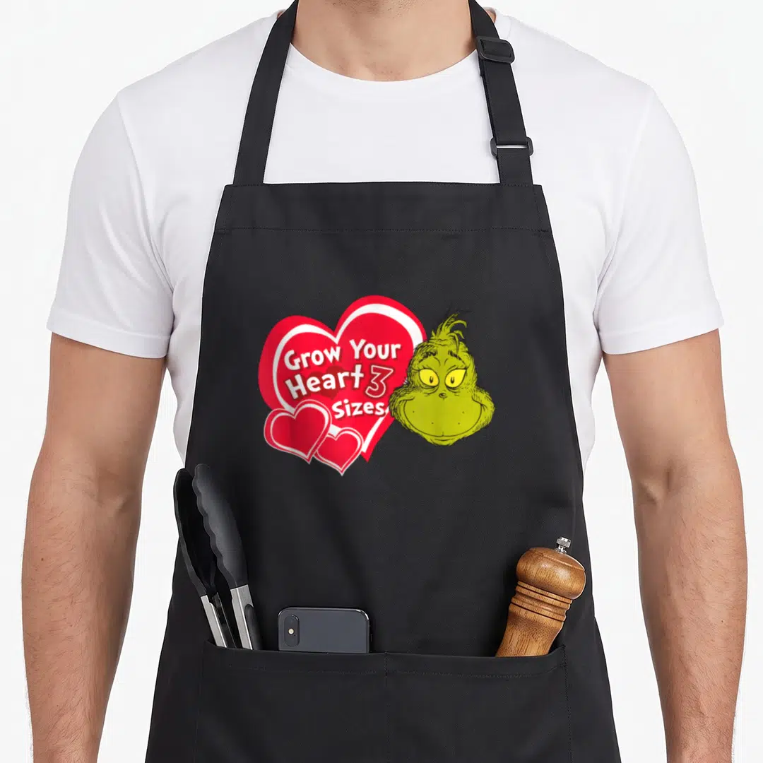 Grow novelty apron - Grinch head next to red heart reading Grow Your Heart 3 Sizes — shown on a black cotton canvas apron against a white background