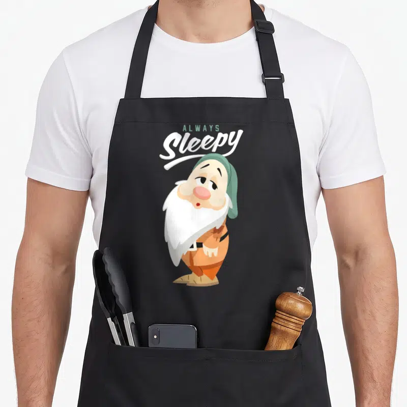 Seven snow white apron - Always Sleepy text above cartoon dwarf in green hat and orange tunic — shown on a black cotton canvas apron against a white background