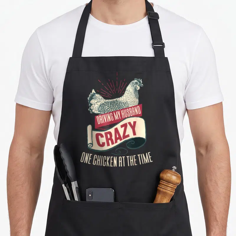 Cool husband driving aprons - Retro chicken illustration with red and beige banners reading DRIVING MY HUSBAND CRAZY — shown on a black cotton canvas apron against a white background