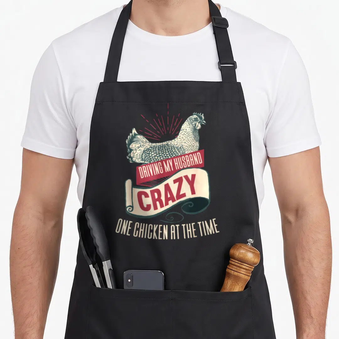 Cool husband driving aprons - Retro chicken illustration with red and beige banners reading DRIVING MY HUSBAND CRAZY — shown on a black cotton canvas apron against a white background