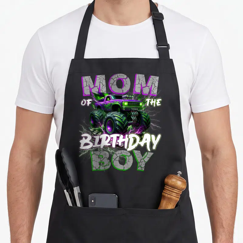 Monster family truck apron - Purple and green monster truck with lightning and MOM OF THE BIRTHDAY BOY text — shown on a black cotton canvas apron against a white background