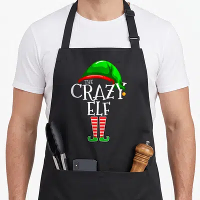 Retro family christmas elf apron - THE CRAZY ELF text between green hat and striped socks — shown on a black cotton canvas apron against a white background