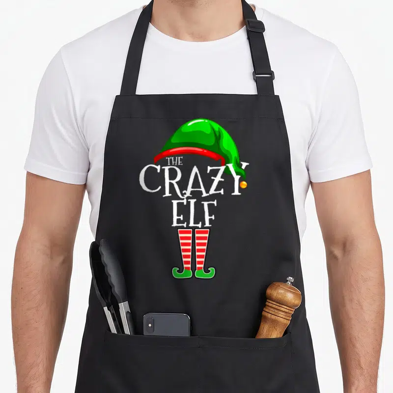 Retro family christmas elf apron - THE CRAZY ELF text between green hat and striped socks — shown on a black cotton canvas apron against a white background