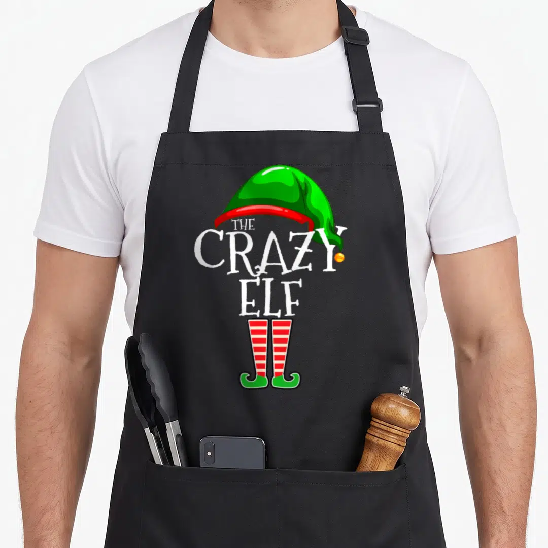Retro family christmas elf apron - THE CRAZY ELF text between green hat and striped socks — shown on a black cotton canvas apron against a white background