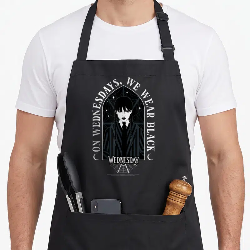 Wednesday goth apron - On Wednesdays We Wear Black text over gothic arch with woman portrait and spiderweb — shown on a black cotton canvas apron against a white background